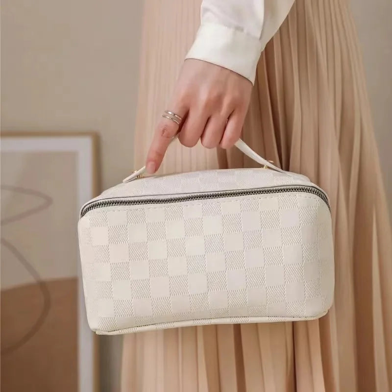 White toiletry bag for women, Fancy material and look, Metal zipper, Front view of bag, Handle, Women's' hand holding it by handle, Bag hanging on side of stylish woman, Neutral colors, Lifestyle image